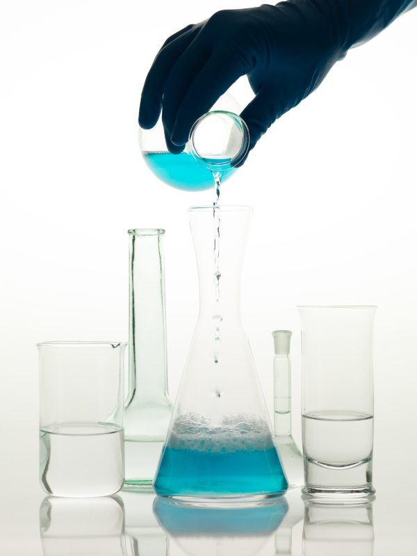frontal view of different types of glass containers used in the laboratory, filled with transparent colorless liquid and one in which someone is pouring a blue liquid